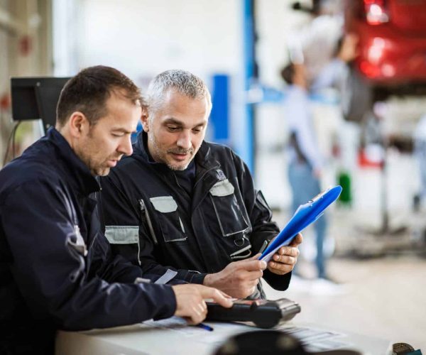 Mid adult auto mechanics working in a repair shop. Auto mechanics cooperating while going through diagnostic list in a repair shop. Focus is on smiling man.